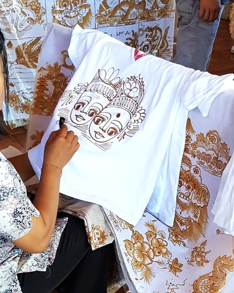 Mano trazando líneas de reserva sobre una prenda blanca de algodón, rodeada de telas con motivos ya dibujados