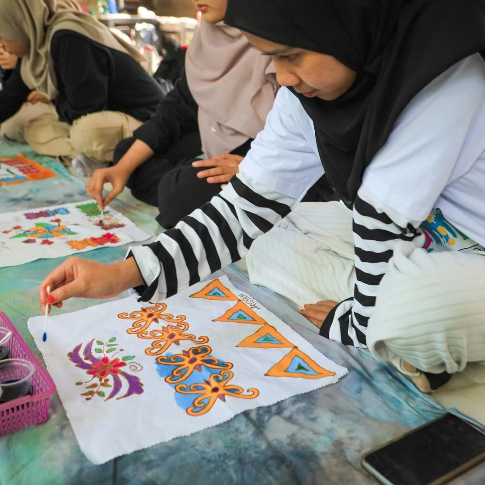 Dos personas coloreando con pincel pequeñas telas de batik cuyos motivos están delimitados por líneas de reserva